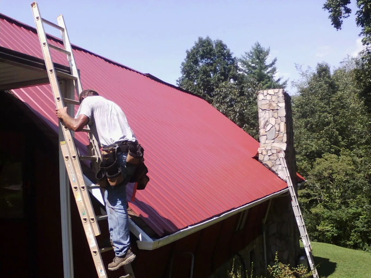 Roofer on a ladder beside a red metal roof and stone chimney — Soffit & Fascia Repair by Roofing Brothers in Snowville